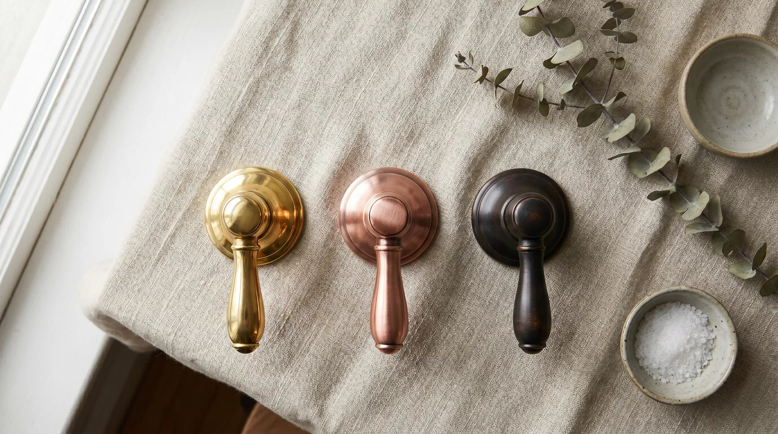 Three metal faucet handles in brass, copper, and oil-rubbed bronze on linen cloth